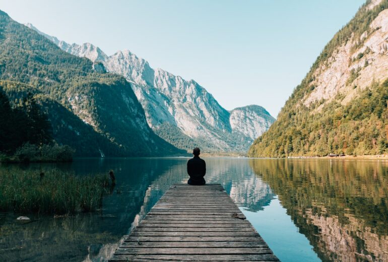 man sitting on a pier at a lake surrounded by mountains